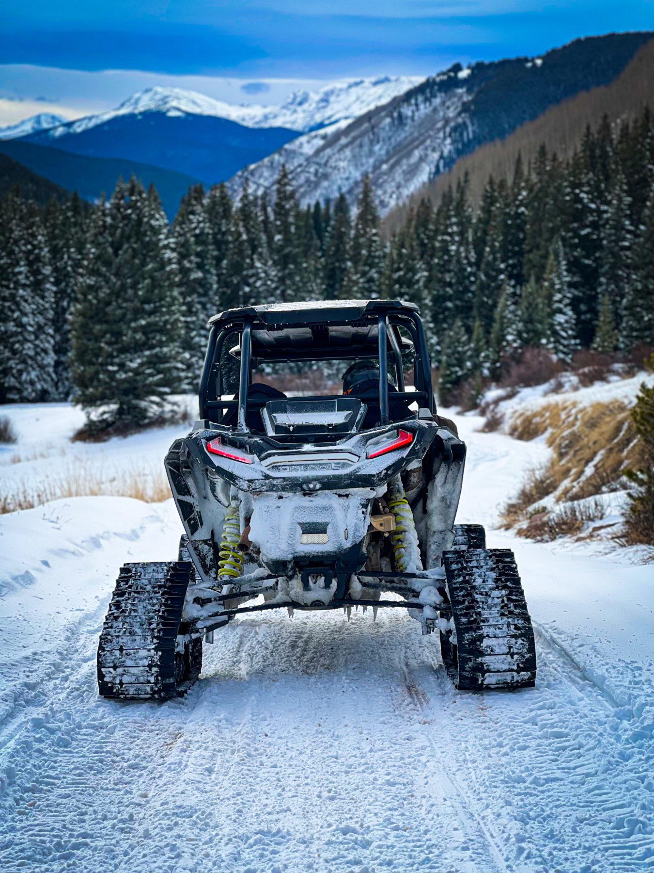 A four-wheeled vehicle with snow tracks is parked on a snow-covered trail in a mountainous, forested area. Snow-covered trees line the path, and mountains under a partly cloudy sky are visible in the background, making it an ideal spot for snowmobile rentals.