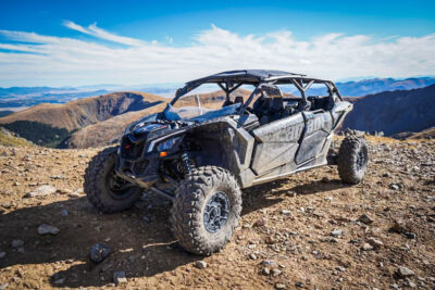 A four-seater all-terrain vehicle (ATV) parked on a rocky mountain trail with a scenic view of mountains and a partly cloudy blue sky in the background. The ATV appears rugged with large, knobby tires and a roll cage.