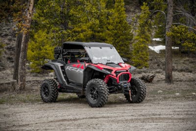 A black and red 2024 Polaris RZR 1000 with large tires is parked on a dirt path in a forested area, surrounded by pine trees and patches of snow in the background.