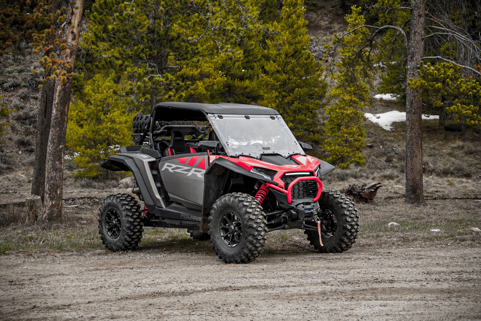 A black and red 2024 Polaris RZR 1000 with large tires is parked on a dirt path in a forested area, surrounded by pine trees and patches of snow in the background.