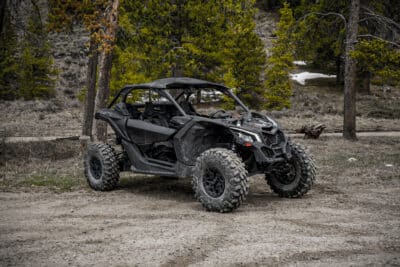 A black 2024 Polaris RZR 1000 XP2 Ultimate off-road utility vehicle with large rugged tires is parked on a dirt path in a forested area, surrounded by trees and patches of snow in the background.