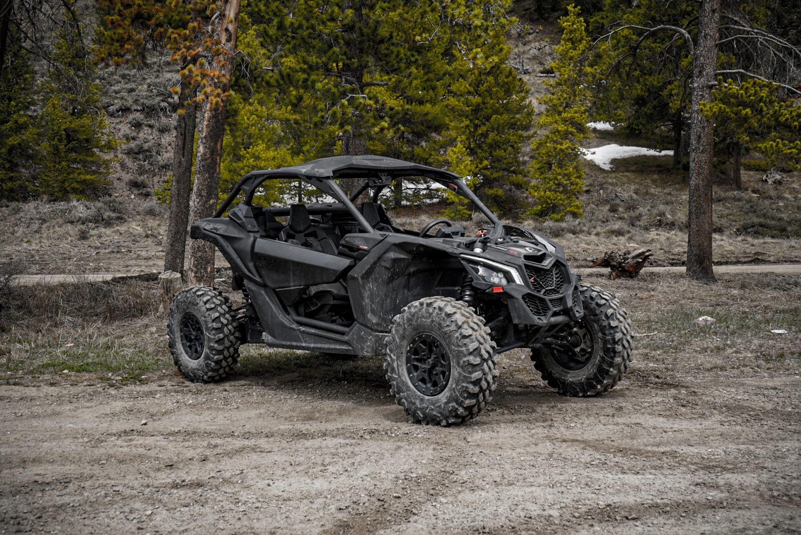 A black 2024 Polaris RZR 1000 XP2 Ultimate off-road utility vehicle with large rugged tires is parked on a dirt path in a forested area, surrounded by trees and patches of snow in the background.
