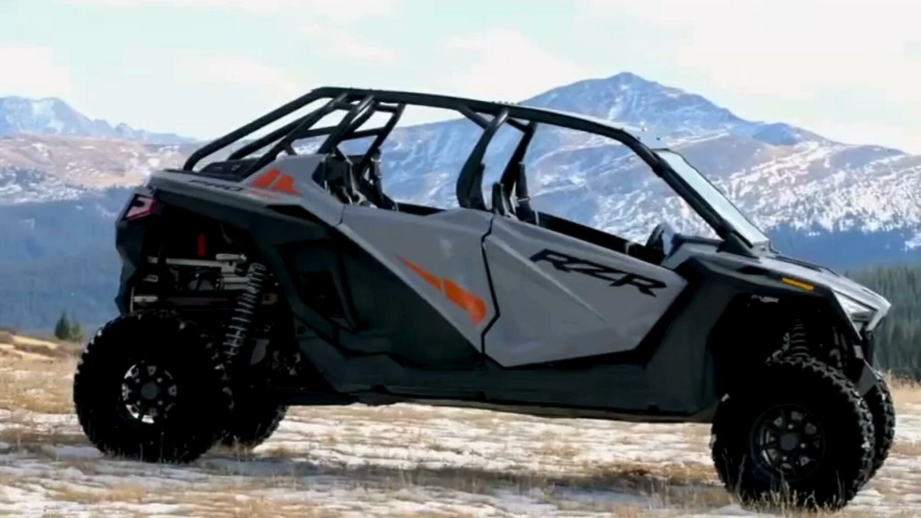 A gray off-road vehicle with a roll cage is parked on a snowy field. Mountains with snow-capped peaks are in the background under a clear sky.