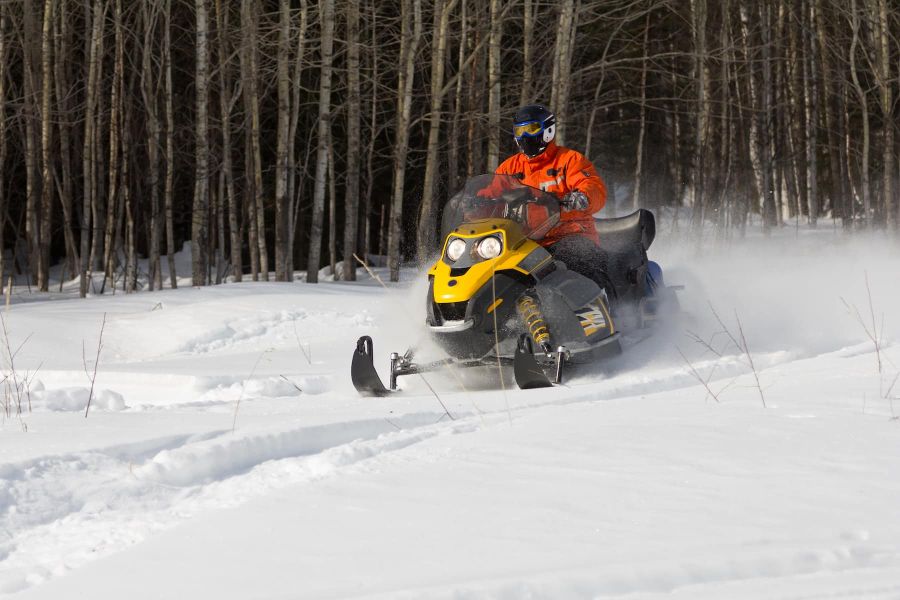 A person in an orange jacket and helmet is riding a yellow snowmobile through a snow-covered forest in Summit County. Snow is kicked up behind the snowmobile, and leafless trees are visible in the background.