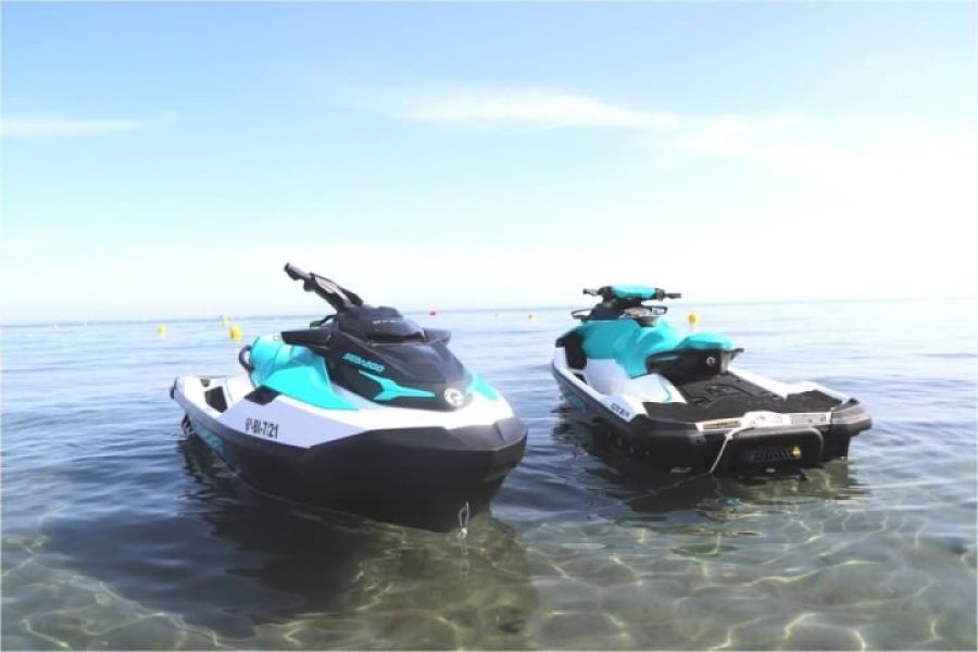 Two jet skis are floating on a calm, clear body of water in Summit County. They are both white with teal accents and facing slightly different directions. The sky above is clear and blue, with some distance between the shore and the horizon, ideal conditions for jet ski rentals near Breckenridge.