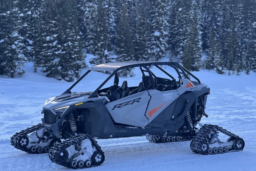A sleek off-road vehicle with tank-like tracks instead of wheels is parked in a snowy landscape, surrounded by dense evergreen trees. The vehicle, reminiscent of the ones you'd find at snowmobile rentals, features a futuristic design with "RZR" branding on the side.
