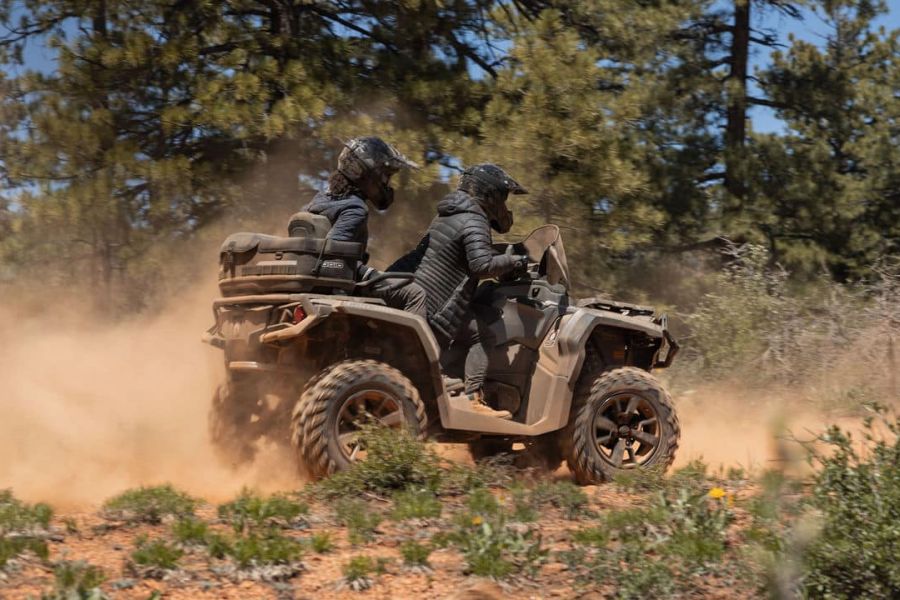Two people in helmets and protective gear ride a beige ATV through a dusty off-road trail surrounded by trees and sparse vegetation. The UTV kicks up a cloud of dust as it moves, creating a dynamic and adventurous scene under the clear Breckenridge sky.