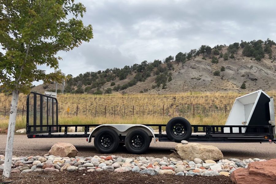 A flatbed trailer carrying a large white object, possibly related to snowmobile rentals, is parked on a gravel area next to a road. The trailer is attached to a silver vehicle. In the background, there are rocks, a grassy hill, and shrubs. A tree is in the foreground on the left side under the cloudy sky.