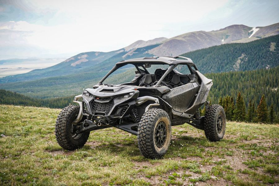 A rugged black off-road vehicle with large tires is parked on grassy terrain, overlooking a scenic mountain landscape with green forests and rolling hills in the background under a cloudy sky.